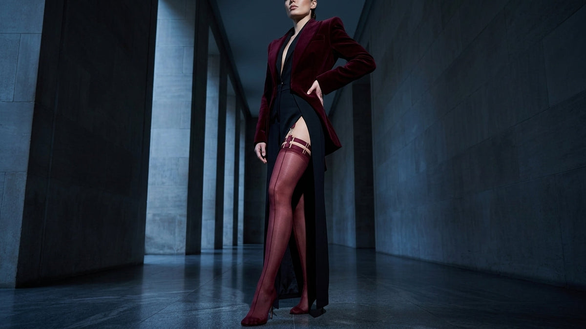 Full-length shot in a columned corridor, burgundy garter stockings with visible strap hardware worn over a black slit dress under a velvet blazer, structured silhouette