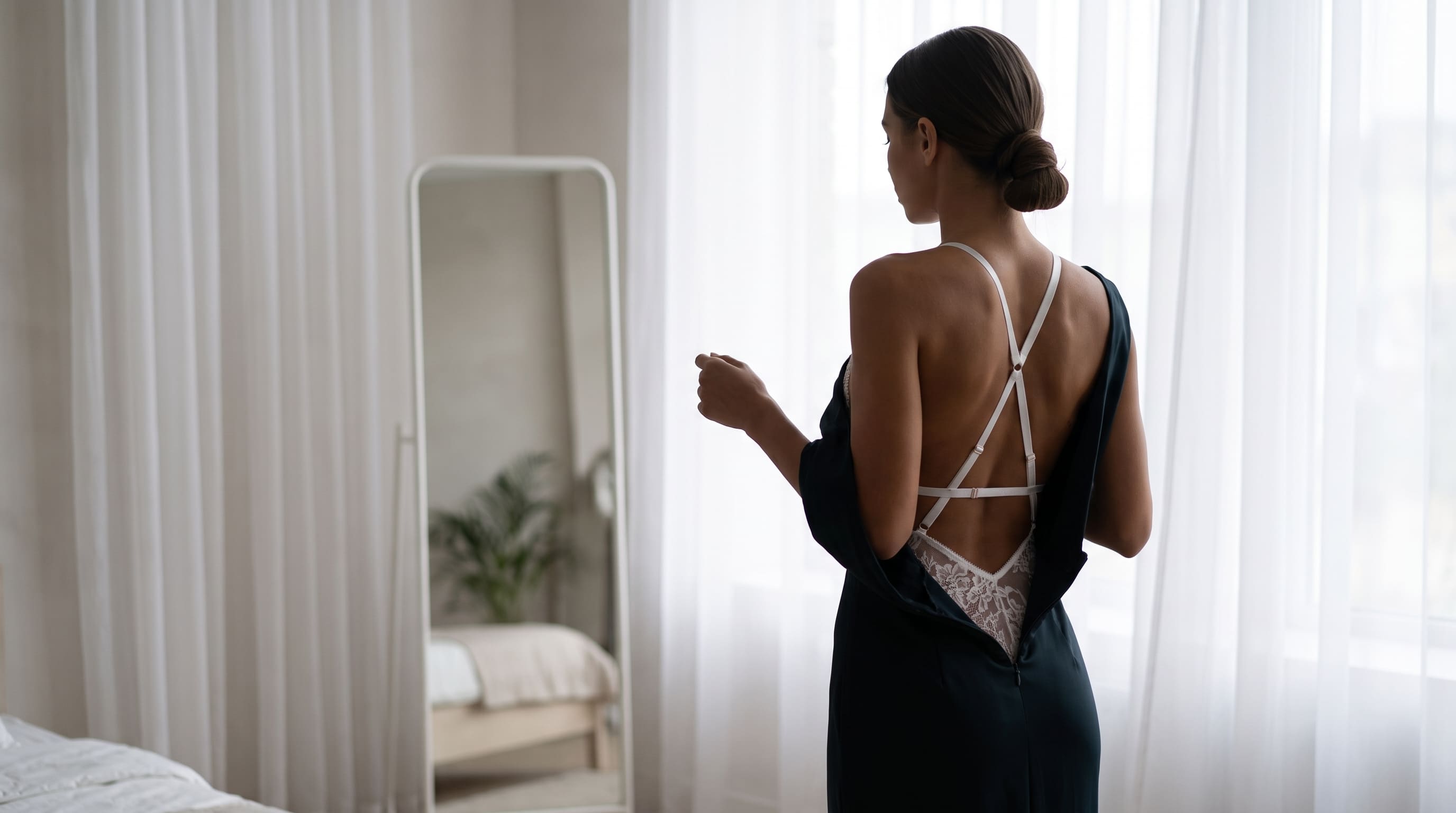 A woman stands with her back to the camera in a bright bedroom, wearing a dark navy slip dress with a white crisscross back strap and lace lining. She is positioned in front of sheer white curtains, with a tall floor mirror reflecting the room.