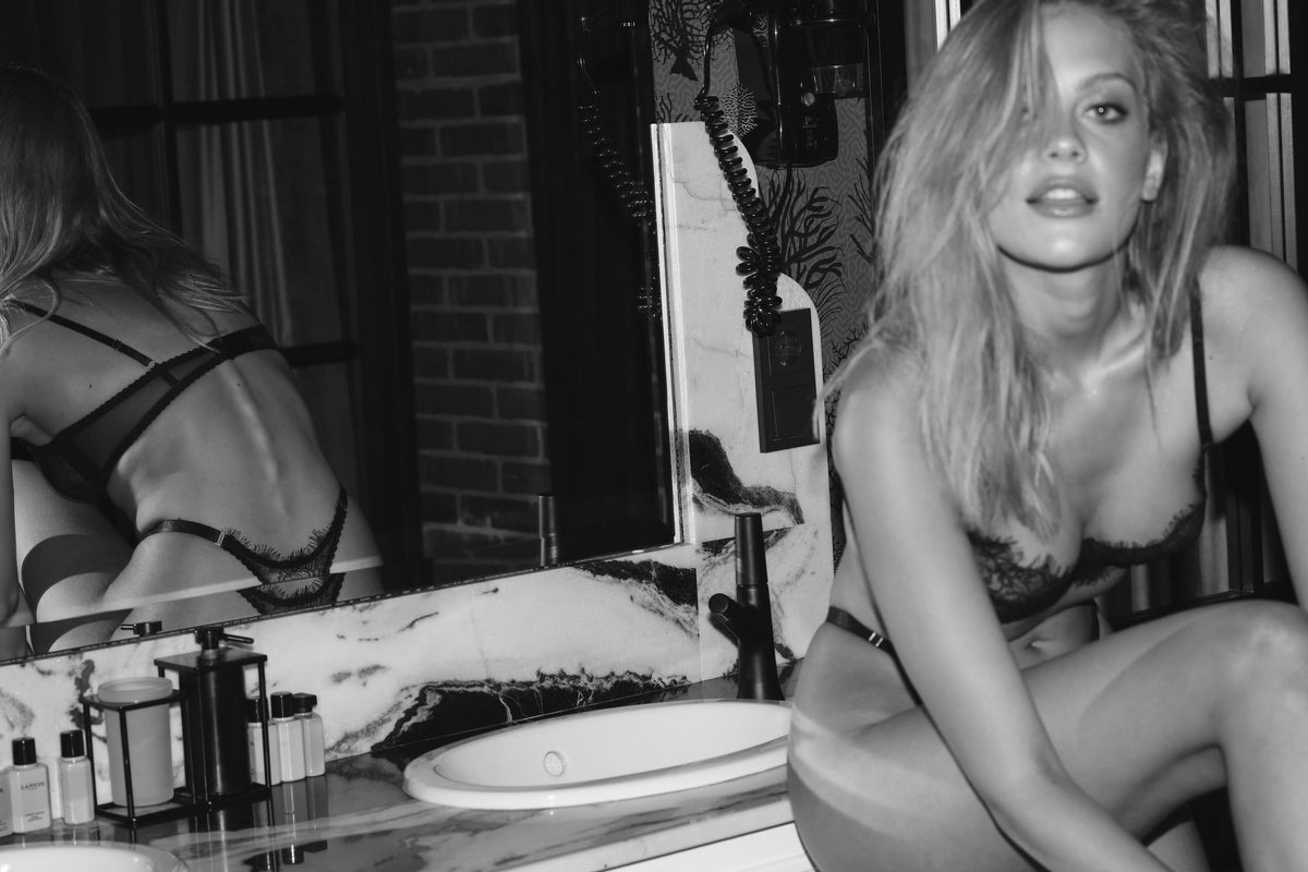 A black and white editorial photograph of a woman in a bathroom, wearing a black lace bra and looking directly at the camera. Her reflection is visible in the mirror on the left, showing the back of the black lace bra. lioness_lingerie